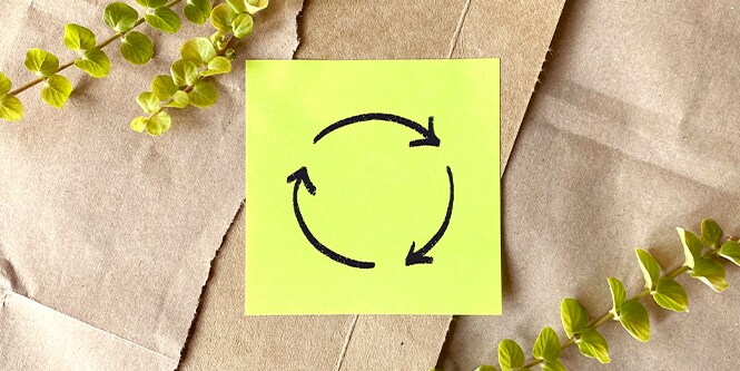 A Yellow Post-it® Note with a recycling icon drawn in white on it, on a craft paper background with plants in the corners.
